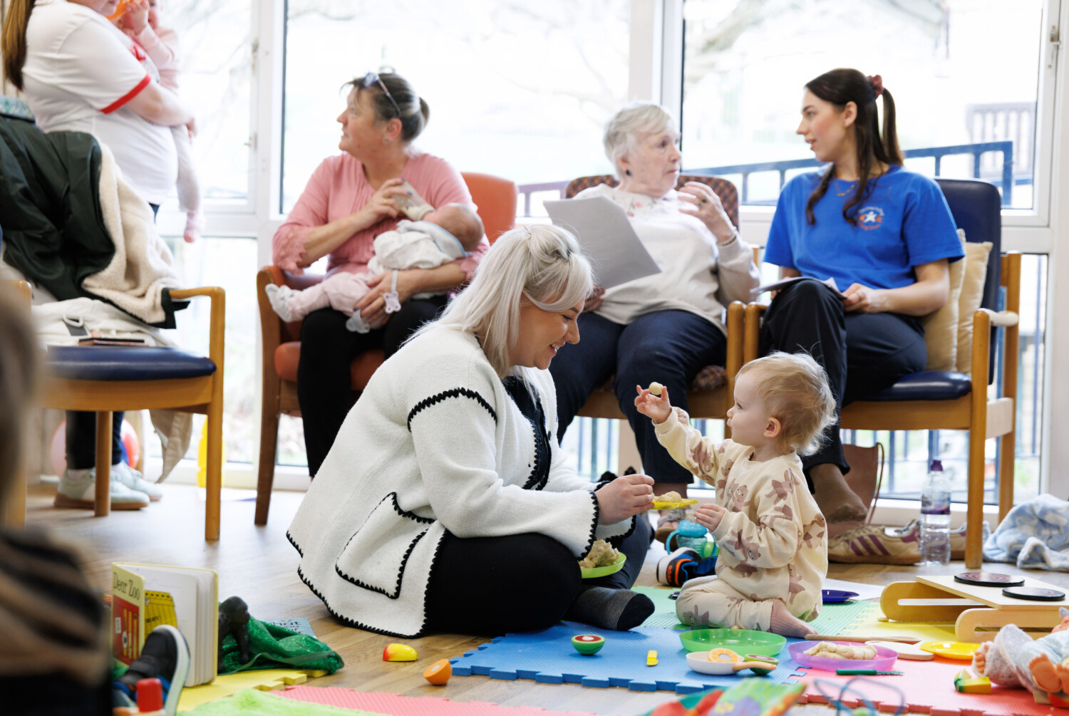 Mother playing with young child and adults sitting on chairs in the background