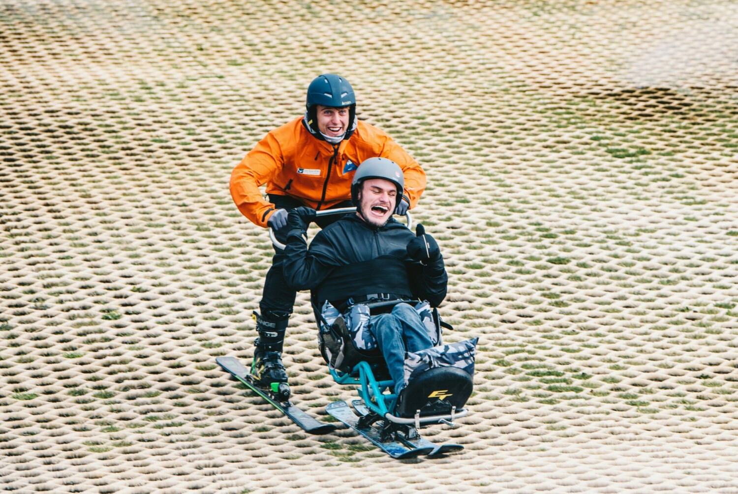 Person, in an orange ski jacket, on skis pushing another person, in a mobility ski, down a dry ski slope