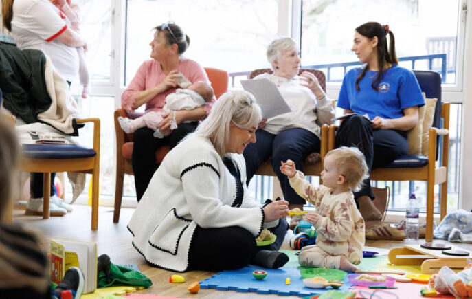 Mother playing with young child and adults sitting on chairs in the background